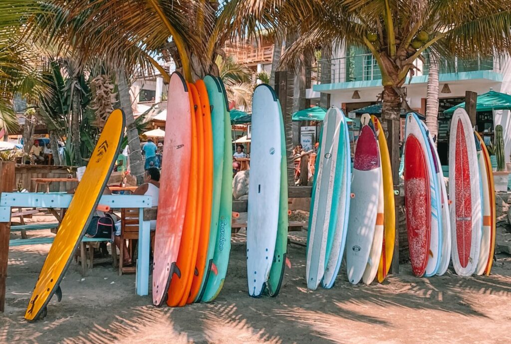 Amplia colección de tablas de surf de colores variados apoyadas en palmeras en una playa con chiringuito al fondo