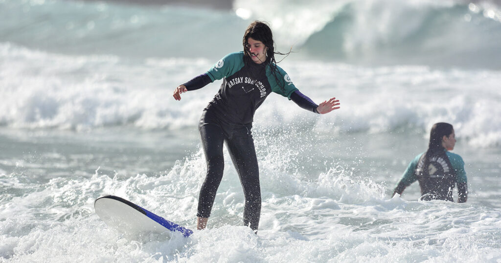 Alumna de pie sobre la tabla surfeando una ola durante una clase de surf en el mar con otra surfista al fondo