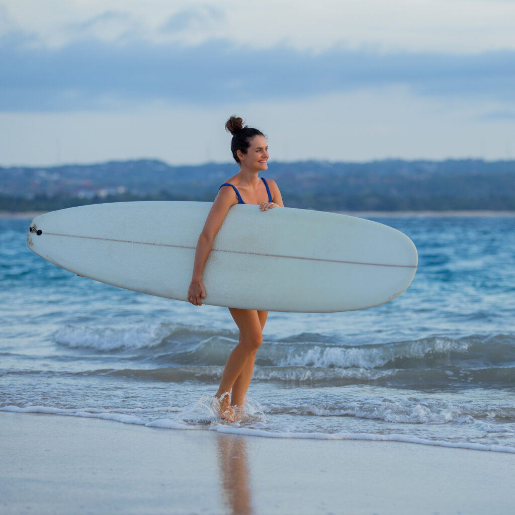 Mujer surfista caminando por la orilla con un longboard blanco bajo el brazo

