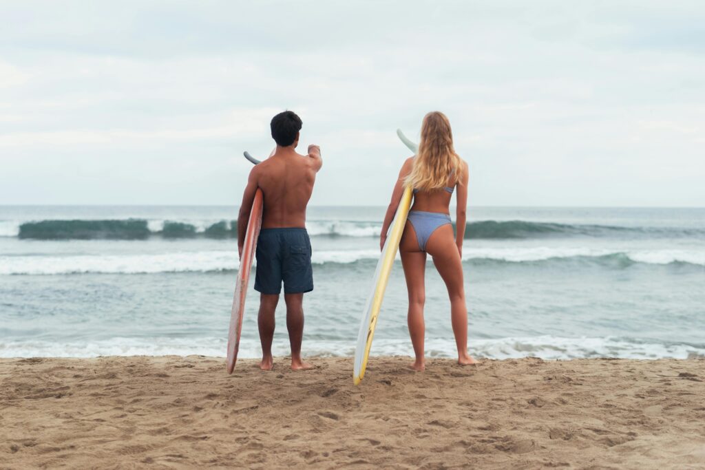 Pareja de surfistas de espaldas observando las olas desde la orilla, cada uno con su tabla de surf