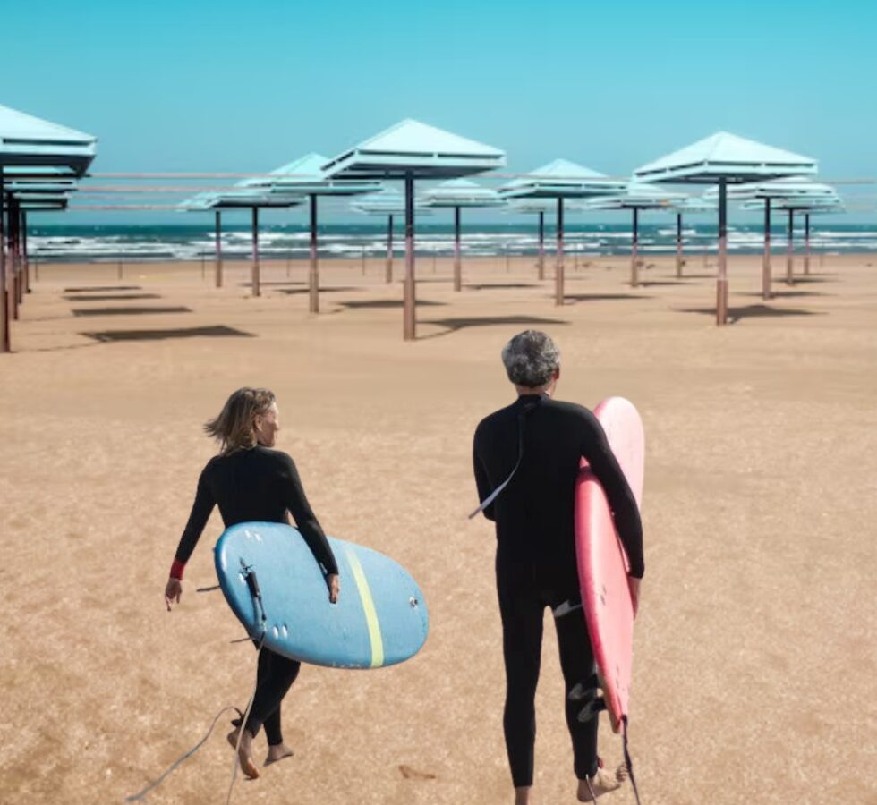 Dos surfistas de espaldas caminando hacia el mar con sus tablas en una playa con sombrillas