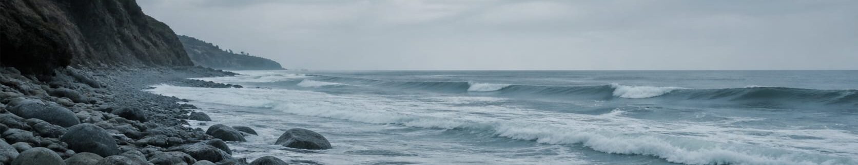 Playa rocosa con olas rompiendo en la orilla bajo un cielo nublado, con un acantilado al fondo